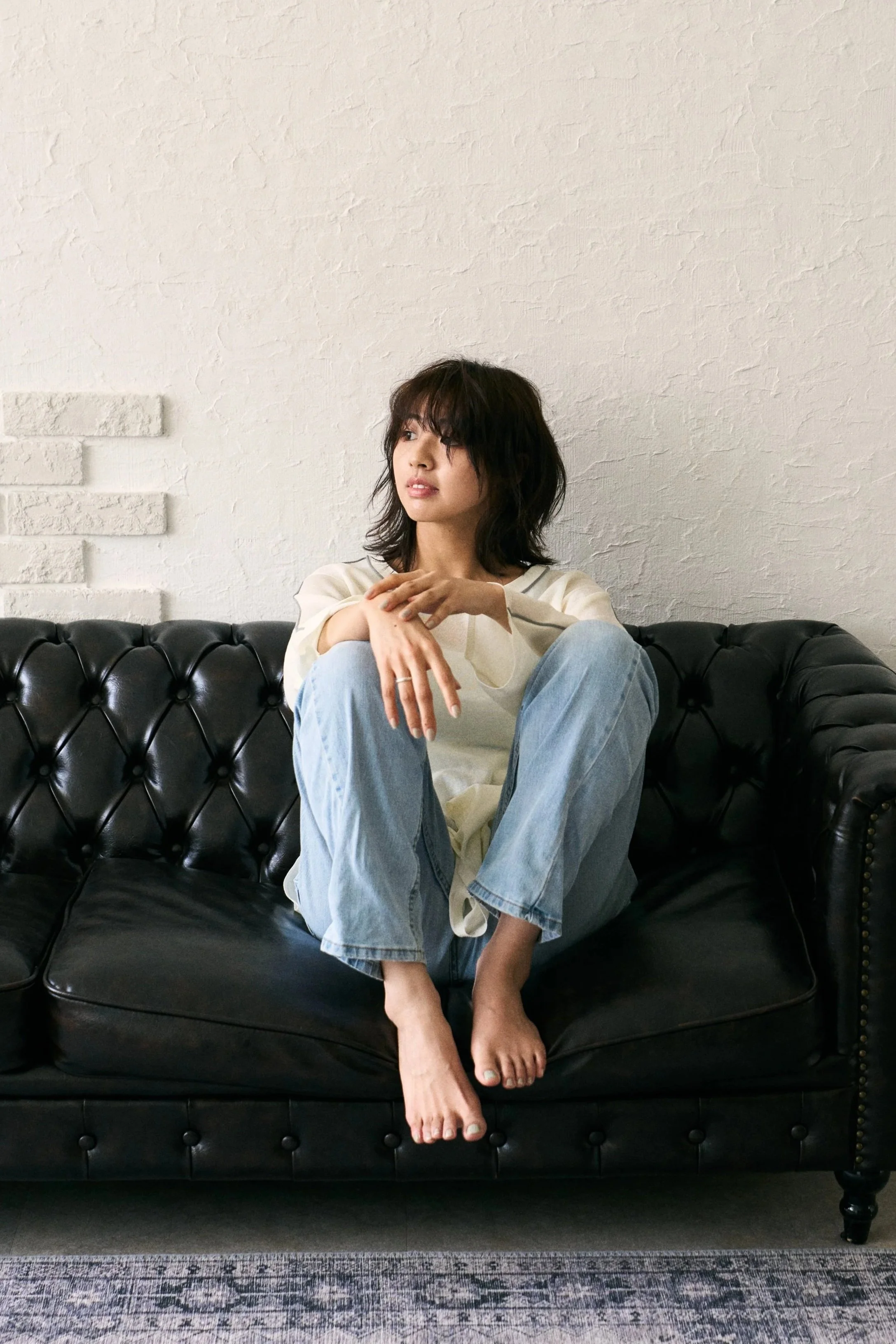 黒い革張りのソファに座る若い女性。白い長袖シャツとジーンズを着ており、壁に向かってやや左を見ている。