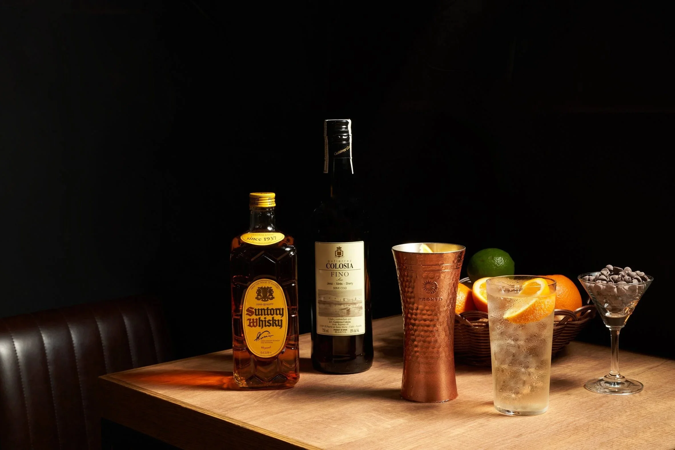 dark background with bottles of alcohol, a copper cup, a glass with lemon and orange slices, and a bowl of blackberries on a wooden table.