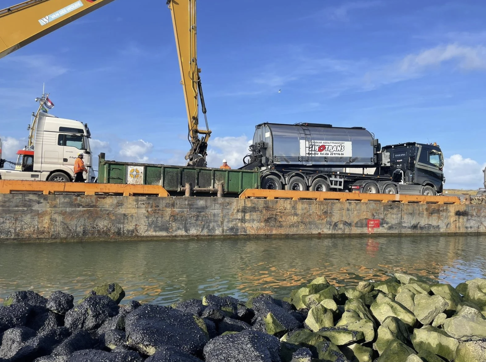 Een boot met een vrachtwagen op het dek die wordt geladen of gelost met een grote grijper door arbeiders, met een waterkant en een stenen oever op de voorgrond en een heldere blauwe lucht op de achtergrond.