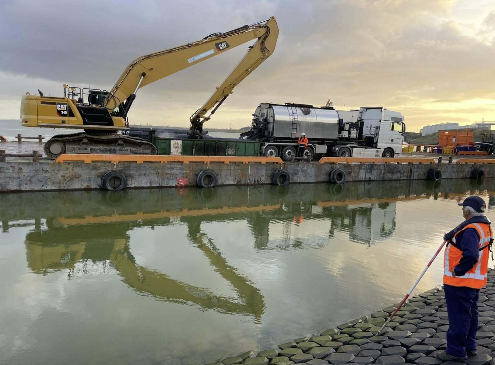Een graafmachine op een ponton die werkzaamheden verricht aan een vrachtauto op het water, terwijl mensen in veiligheidsvesten het water inspecteren.