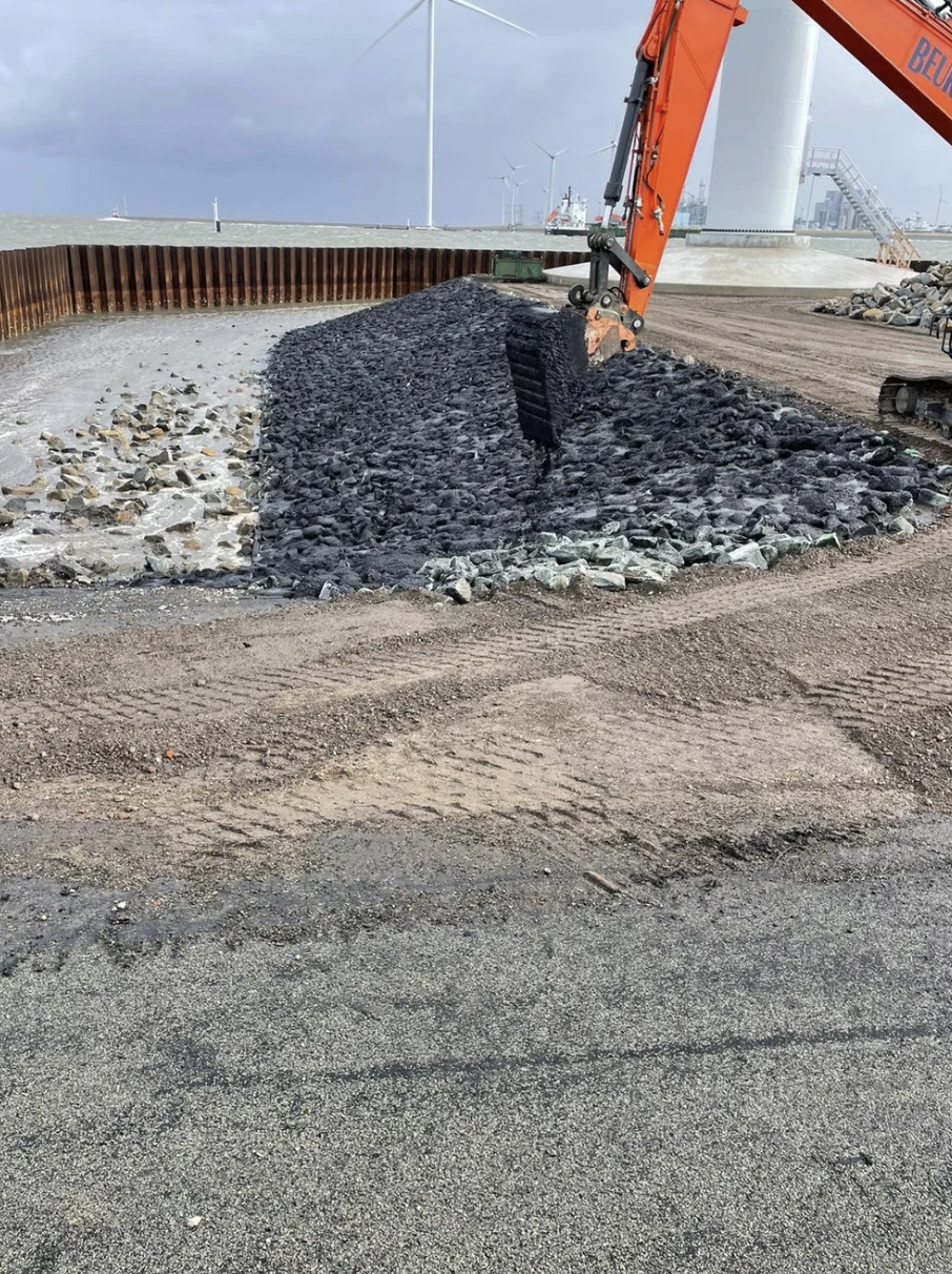 Een bouwput met een graafmachine die zwarte grind of stenen in een gracht of greppel aan het scheppen is, met windturbines op de achtergrond en een gemengd landschap van zand en asfalt.
