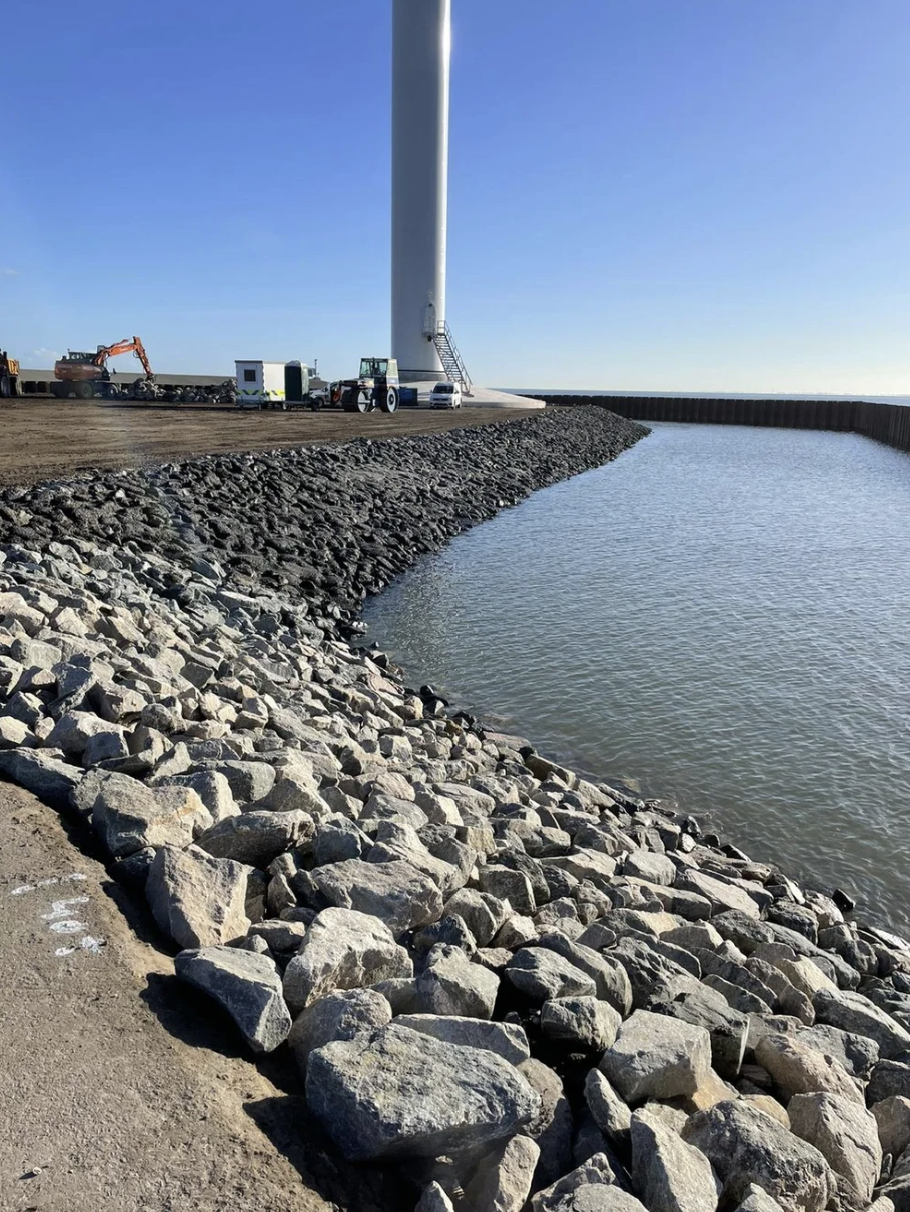 Een haven met een rotsenbaken en een windmolen. Verschillende bouwmachines en voertuigen staan op de kade.