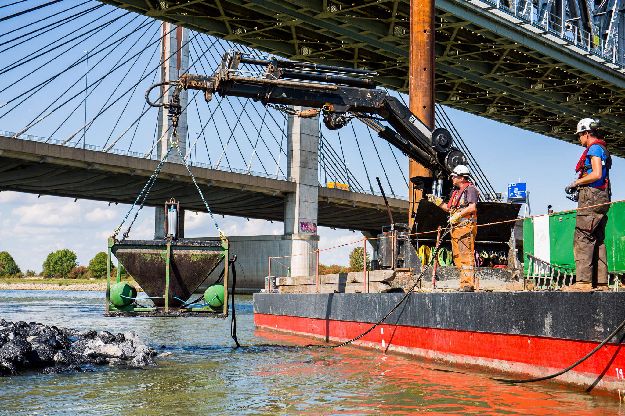 Bouwvakkers werken aan de onderkant van een brug boven een rivier, met een hydraulisch hijsmechanisme die een object omhoog tilt, terwijl zij een fundament of structuur repareren of bouwen.