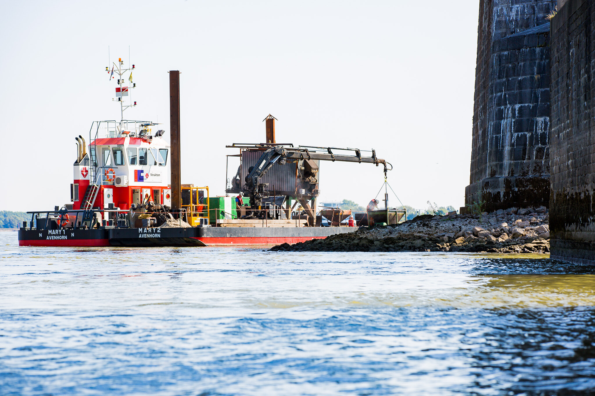 Een sleepboot op een rivier dat een bouwkraan en een walmuur passeert.