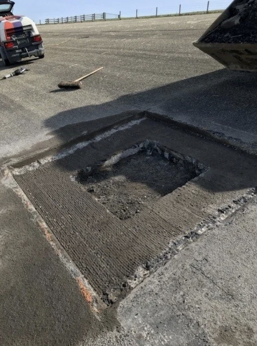 Helft van een ingegraven elektrische leiding of kabel in de stoep, omgeven door ruwe cementrand, met verkeersborden en voertuigen op de achtergrond.