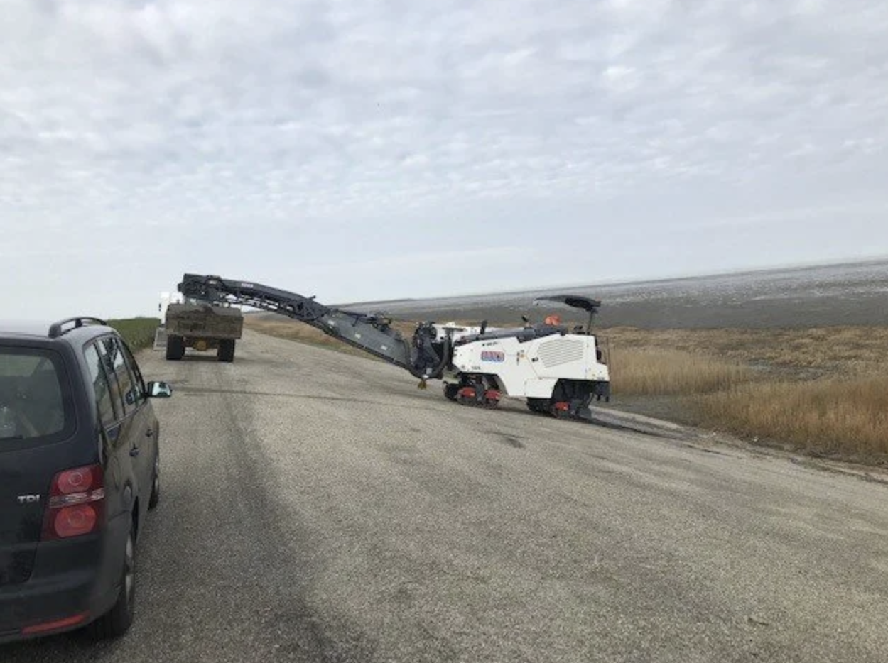 Een graafmachine die aarde wegschept langs een weg in een landelijk gebied met een bewolkte hemel.