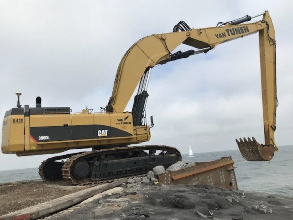 Geleende gele excavator met de tekst 'VAN TUNEN' op de arm, staat op een stenen en zandige aanlegsteiger aan de waterkant met een klein zeilboot op de achtergrond.