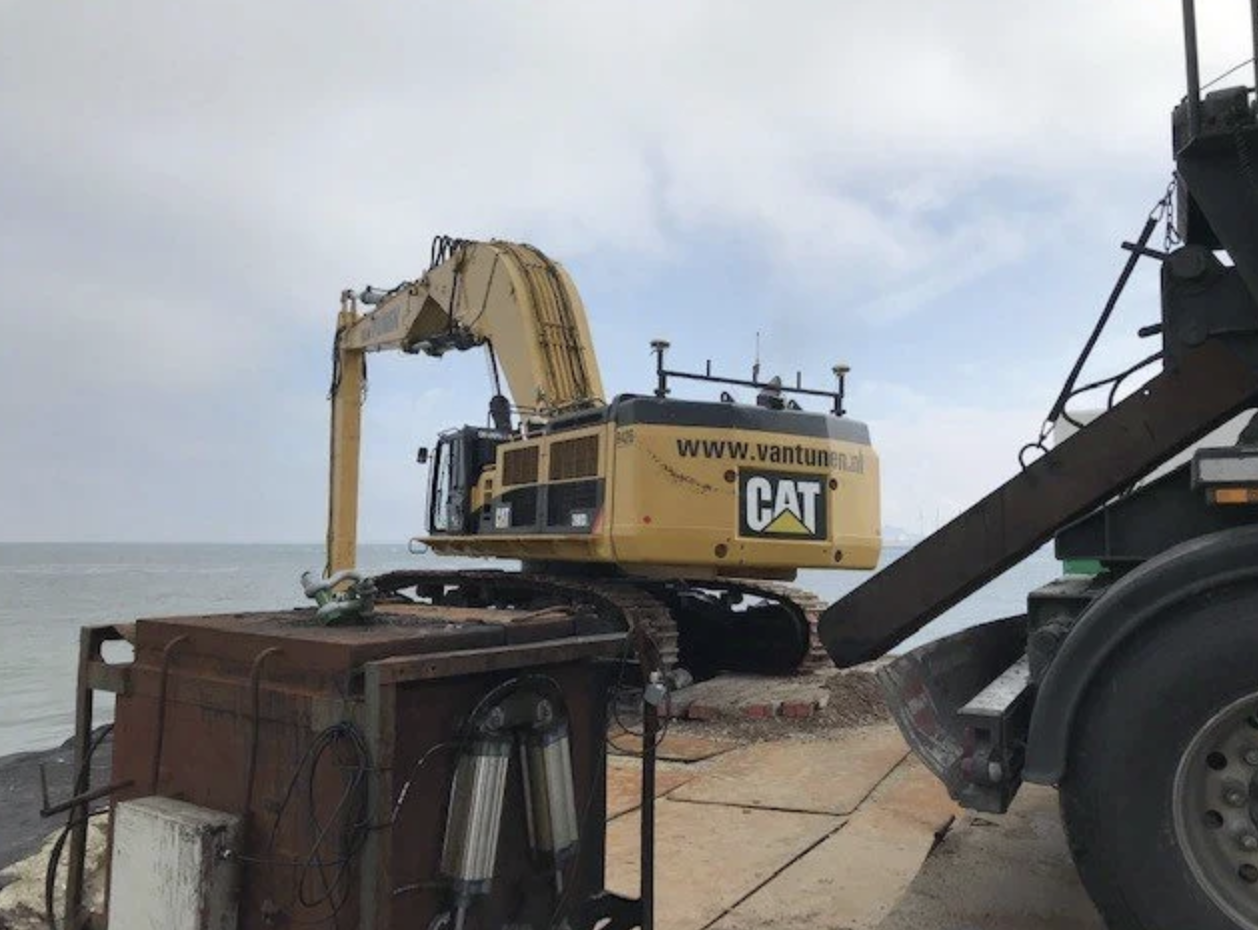 Een gele Caterpillar graafmachine op een strand, met de zee op de achtergrond.