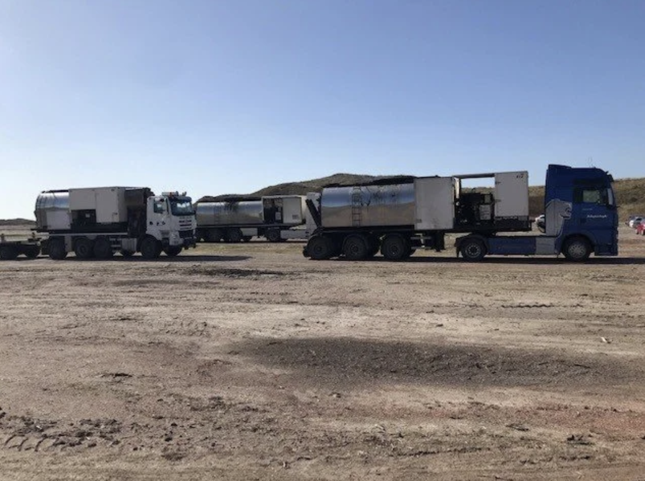 Twee vrachtwagens met tankcontainers staan stil op een stoffige weg, met heuvels op de achtergrond onder een heldere blauwe hemel.