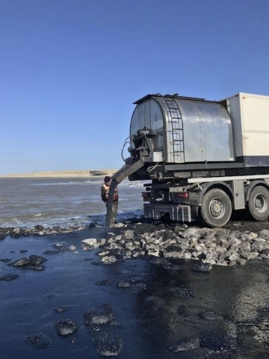 Persoon staat bij een grote machine die zand uit een waterige rivier of meer pompt, met een landschap en de blauwe hemel op de achtergrond.