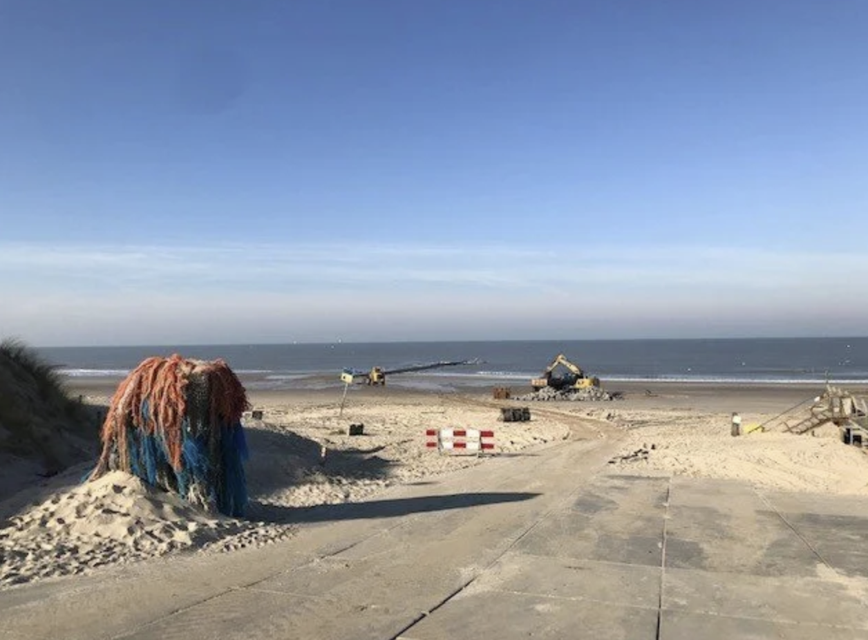 Straatafzetting op het strand met bouwmachine, netten en zandduinen onder een blauwe hemel.