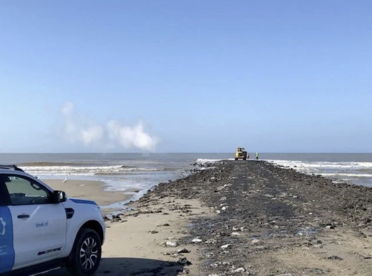 Een strand met een weg van stenen en zand die uitloopt in de zee. Aan de linkerkant een politieauto en op de weg een vrachtwagen en een persoon in een fluorkleurige jas.