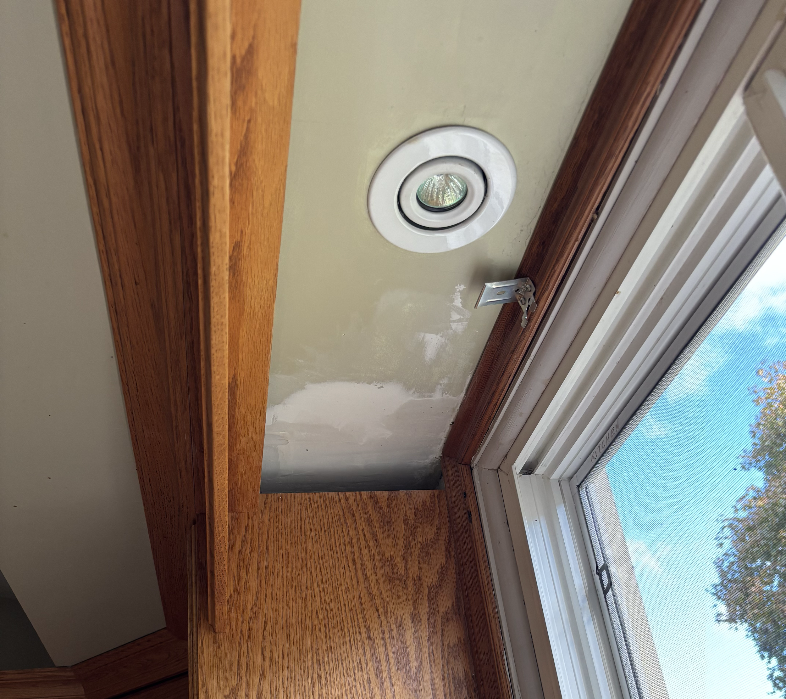Close-up of a ceiling light fixture near a window, with wooden trim on the ceiling and window frame, and a view of trees and blue sky outside.