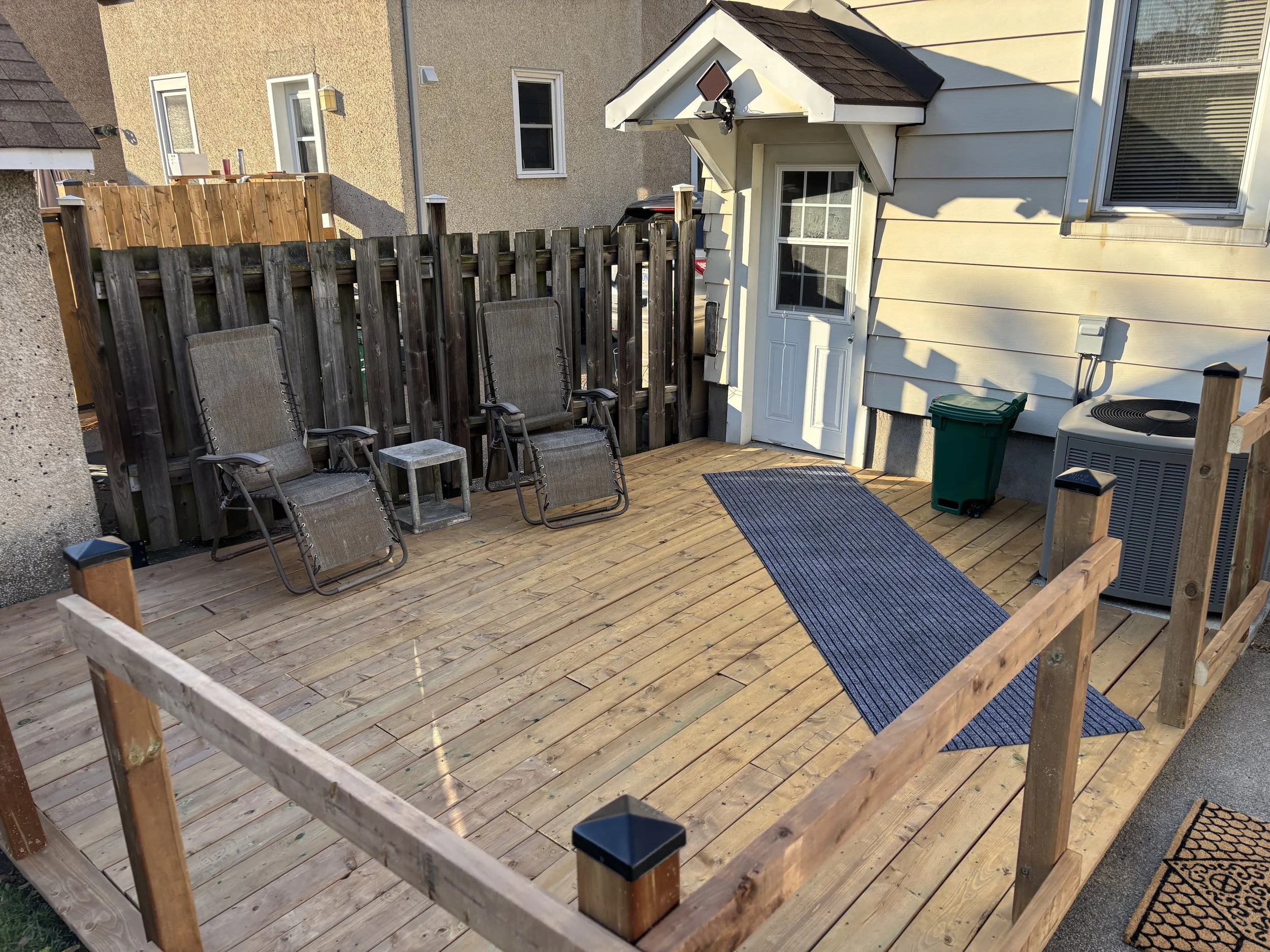 A small backyard deck with wooden flooring, three patio chairs, a small table, a gray and black outdoor storage box, a green trash bin, an air conditioning unit, and a rear door with windows. There is a wooden fence in the background, and part of a neighboring home is visible. The deck is enclosed by a new wooden railing.