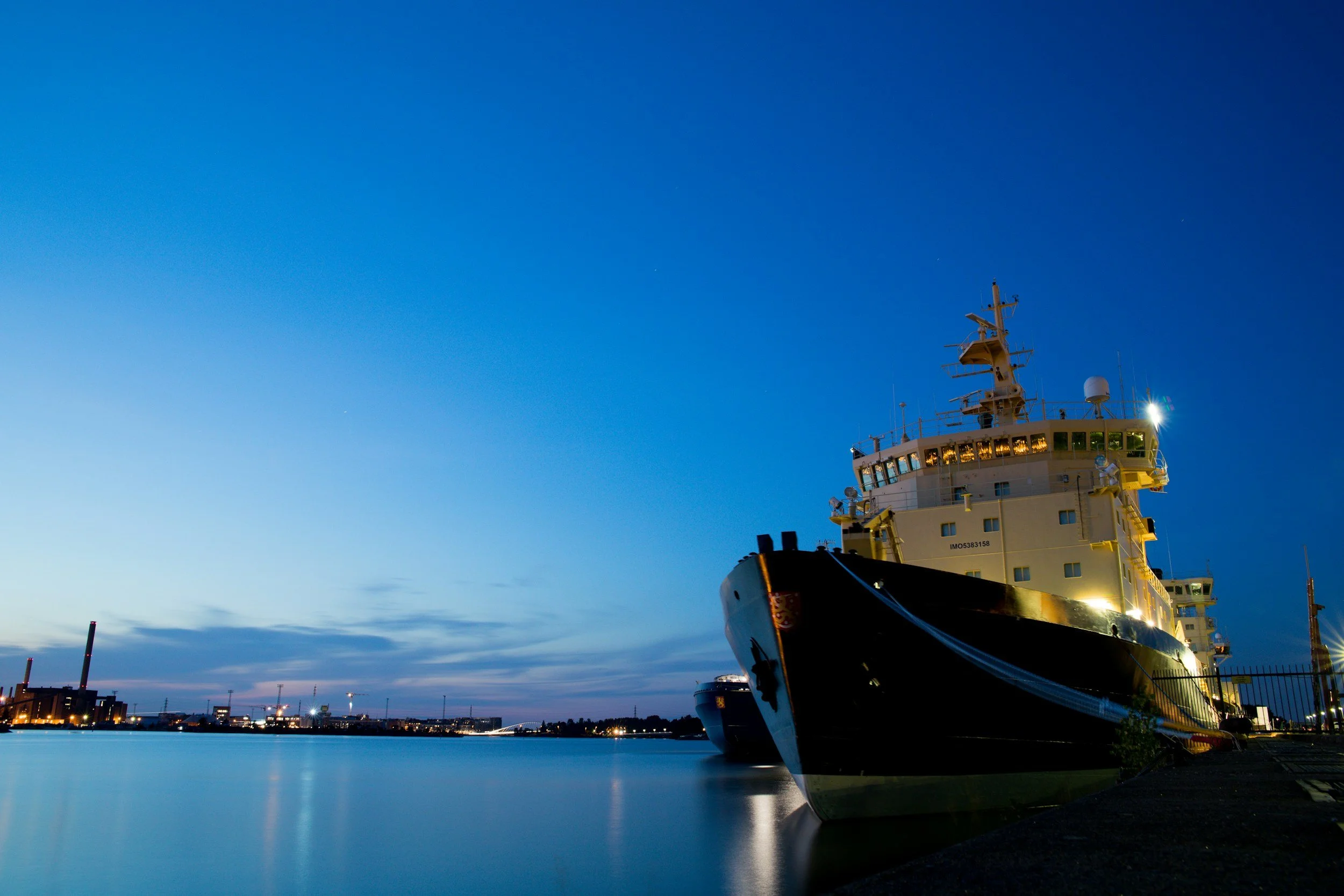 A large cruise ship docked at a harbor during twilight with city lights in the background.
