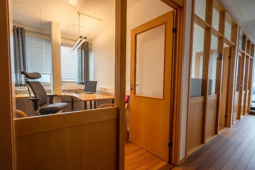 Office cubicles with wooden partitions, door open to an office with a desk, office chair, and computer, sunlight coming through windows with blinds.