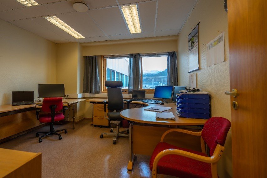 An office with a desk, multiple monitors, and chairs, with a window showing mountains outside.