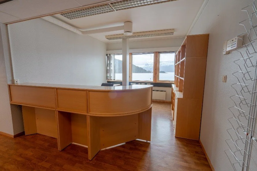Interior of a room with wooden furniture, large window with scenic view of water and mountains, and a white wall with textured surface.