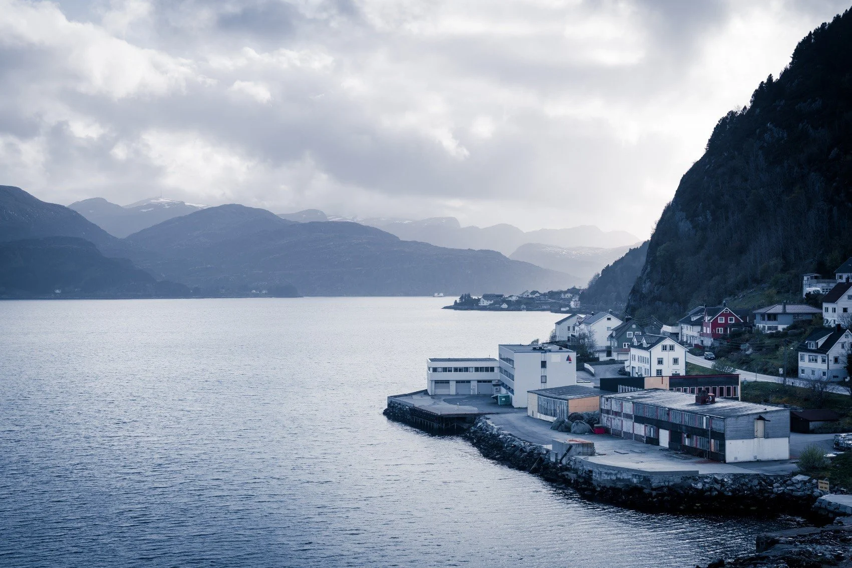 A coastal village by a large body of water, with mountains in the background and a cloudy sky overhead.