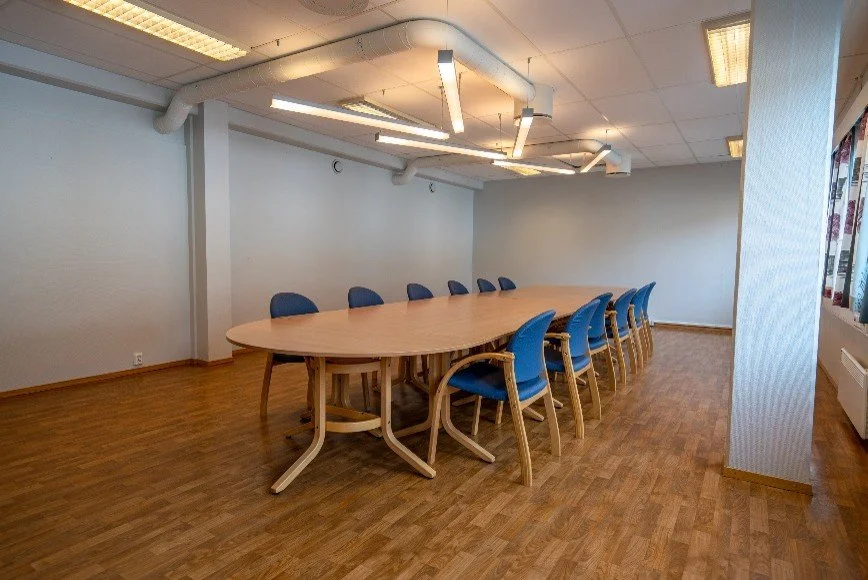 Empty conference room with a long oval wooden table and blue chairs, lit by modern ceiling lights, with wood flooring and plain walls.
