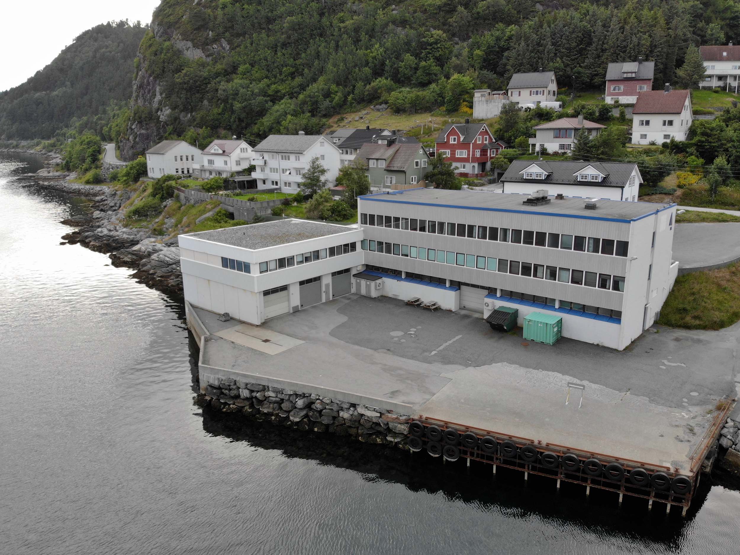 A coastal scene with tiered houses on a hillside overlooking calm water with rocky shoreline, and a large modern white building in the foreground.