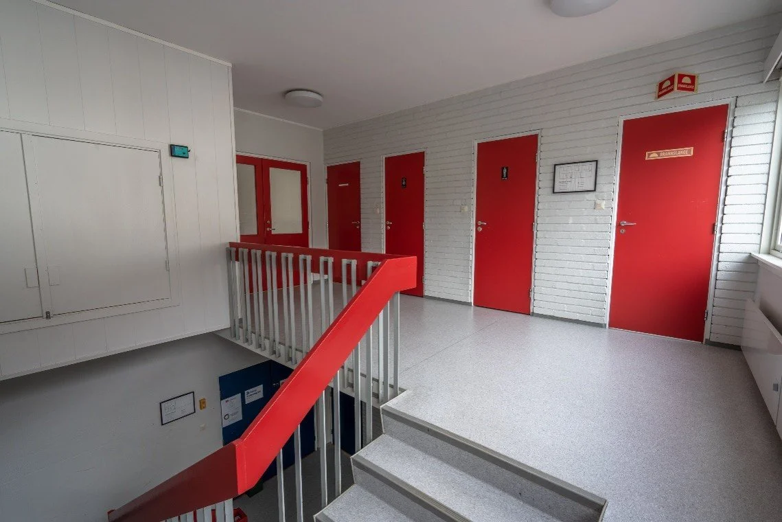 A hallway with white brick walls, four red doors, and a staircase with a red handrail leading down, with fire safety signs and a window on the right.