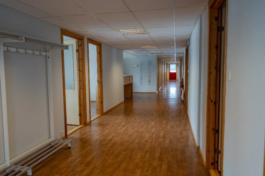 Empty hallway in an office or commercial building with wood flooring, white walls, and wood trim around doors. There is a coat rack on the left side and a small counter on the left side farther down. At the end of the hallway, there is a red door wit
