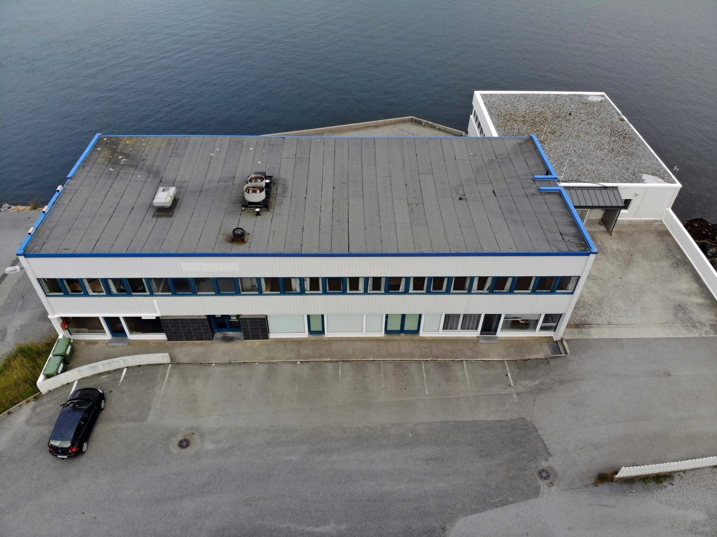 Aerial view of a modern, flat-roofed building with a rooftop parking area beside a large body of water, with a few parked cars on the street.