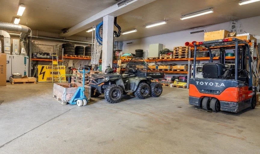 Indoor storage area with a small green utility vehicle, a blue hand truck, and a black and orange forklift. Shelves with supplies and equipment are visible in the background.