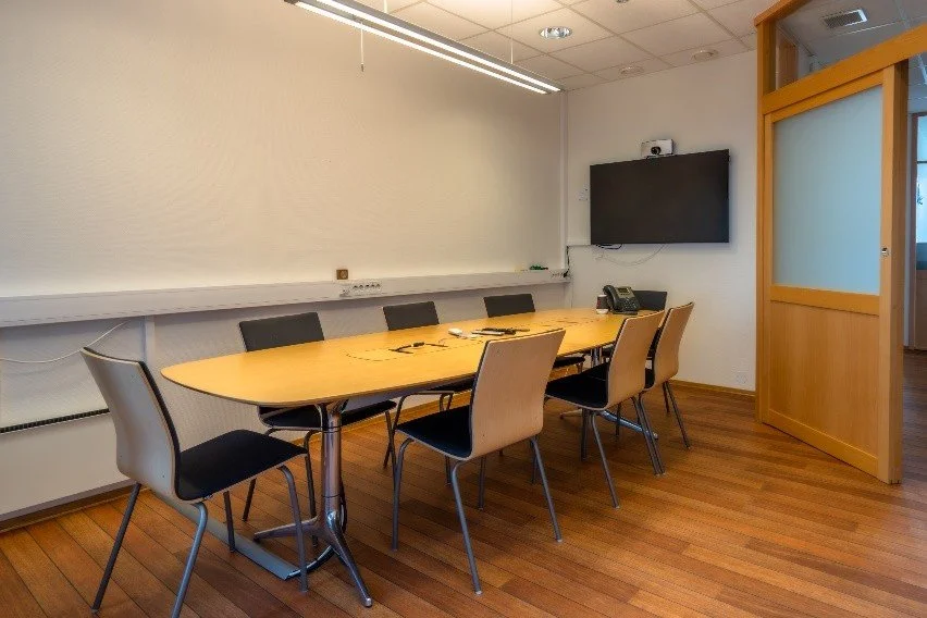 Empty conference room with an oval table, six chairs, a wall-mounted TV, and a frosted glass door.