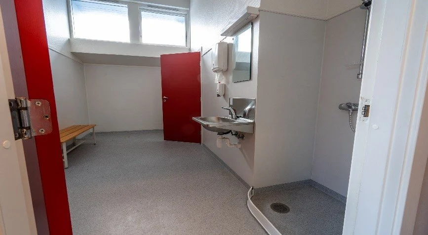 Empty changing room with a bench, a door, a sink, a mirror, and a shower area.