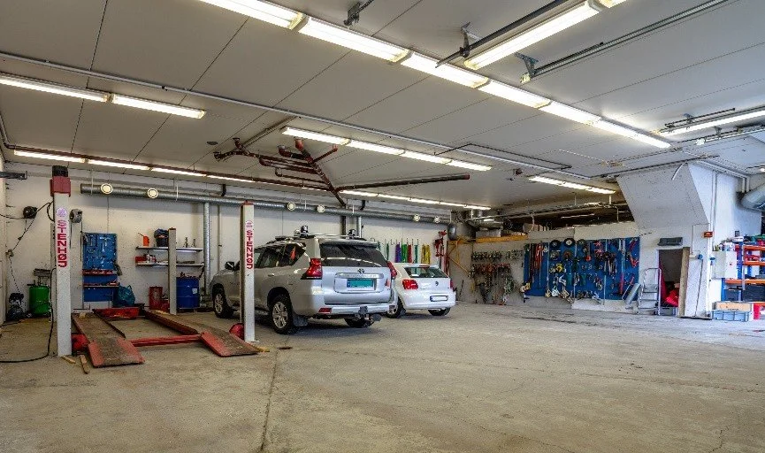 An indoor parking garage with two cars parked on the left, a car lift, and a tool wall on the right, illuminated by ceiling lights.