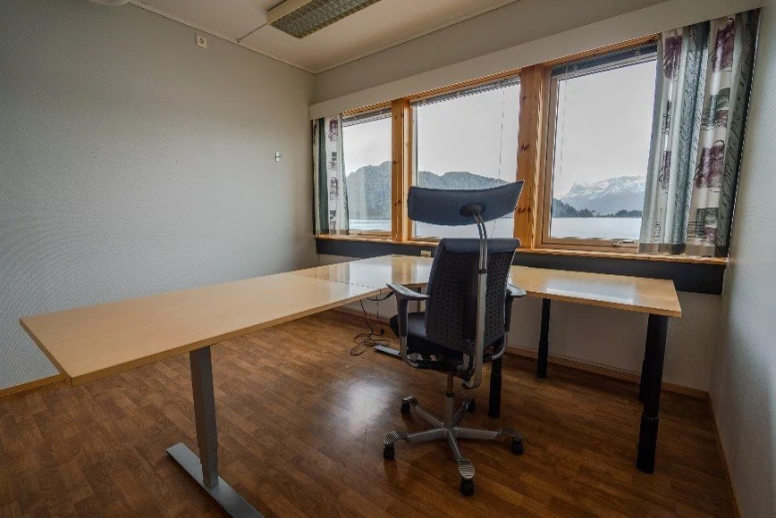 Empty office with a wooden desk, an ergonomic chair, and large windows showing a mountain and lake view.