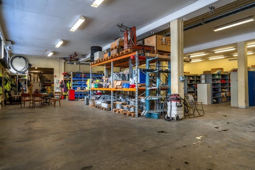 Interior of a warehouse with industrial shelving units filled with boxes and equipment, and a vacuum cleaner on the floor.