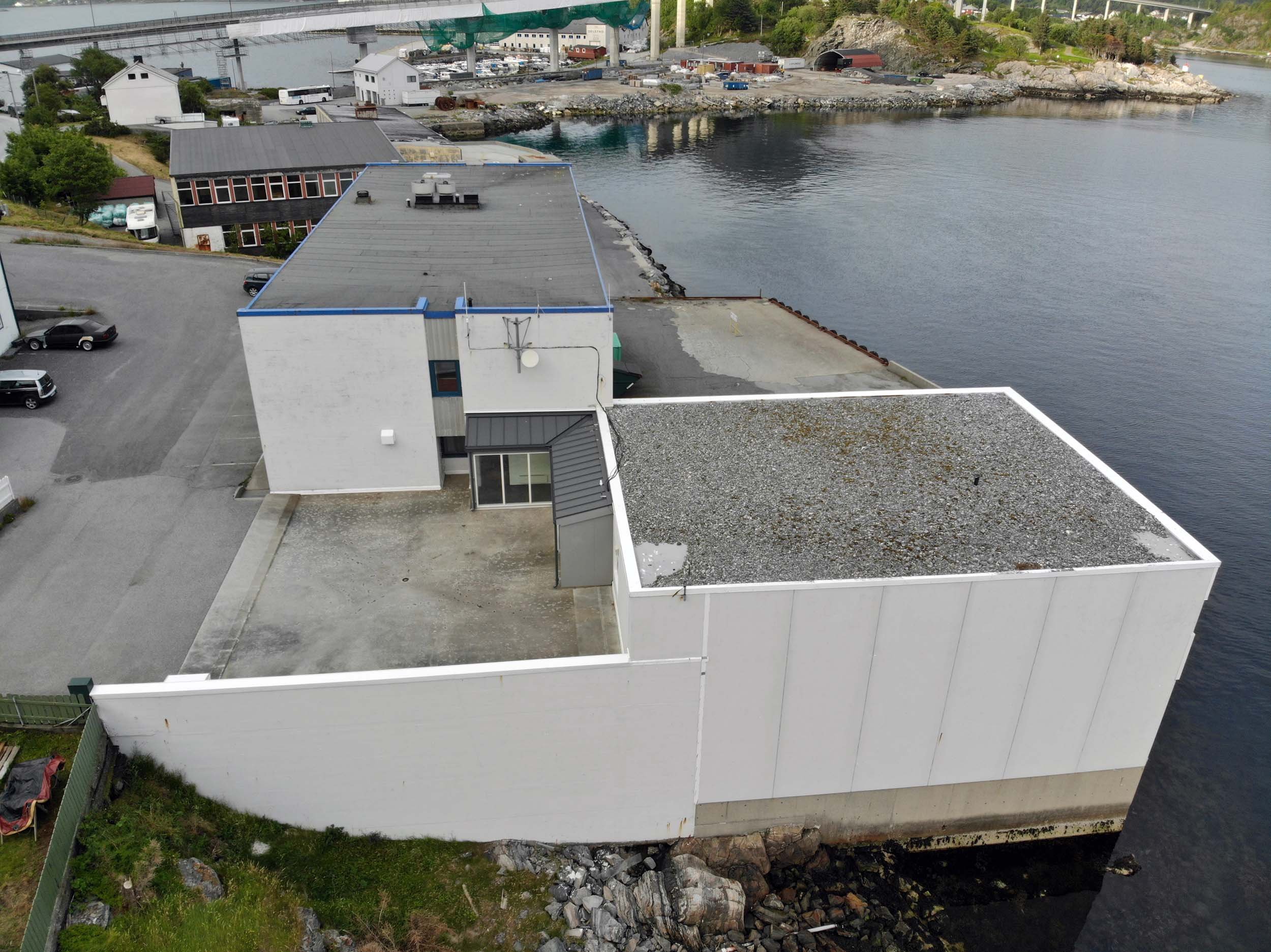 Aerial view of a coastal area with a white building, parking lot, and water. The building has a flat roof and is next to a rocky shoreline with some greenery. There are other industrial buildings and a bridge visible in the distance.