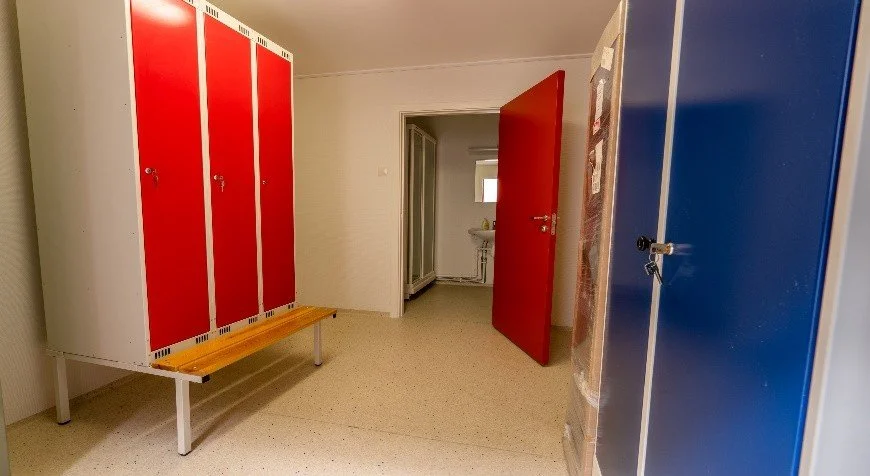 Locker room with red and blue lockers, a wooden bench, and a door leading to a room with a sink.