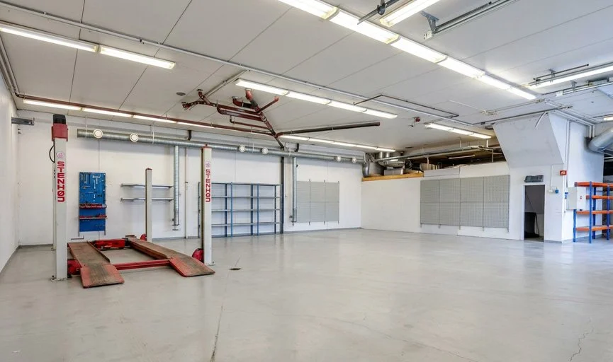 Tomt industri- eller verkstedshall med gulv, veggmontert utstyr og hyller, opplyst med taklys.