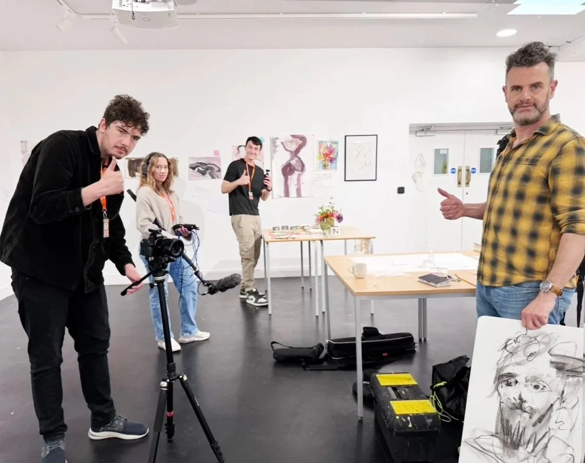 Group of people in art gallery or classroom, one man with a camera, others displaying artwork and giving thumbs up.