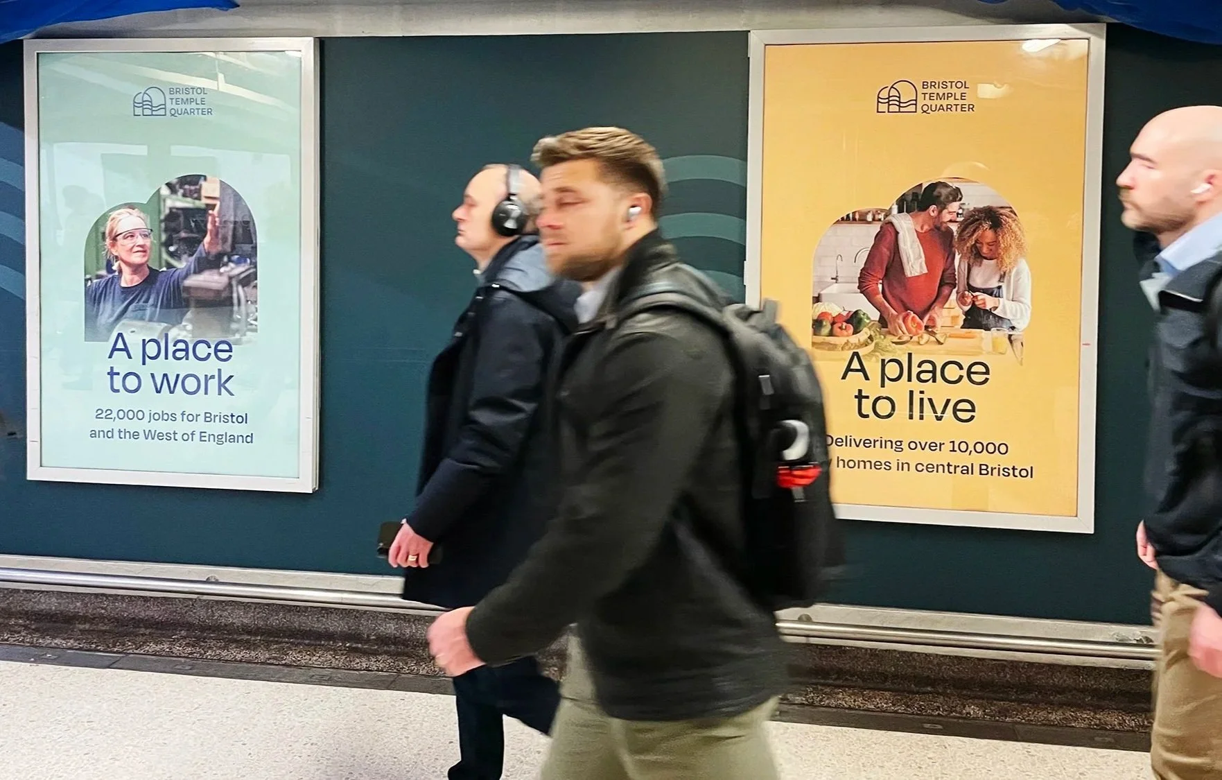 Two posters in an underground passage. The left poster has a woman with glasses taking a photo, with the text "A place to work" and information about jobs in Bristol. The right poster shows a man and a woman cooking together, with the text "A place to live" and information about delivering homes in central Bristol. Several people are walking past the posters.