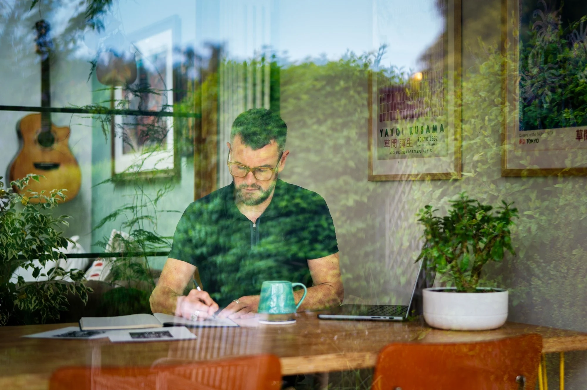 A man with glasses and a beard sits at a wooden table, writing in a notebook, seen through a window with greenery and framed posters inside, including one titled 'YAYOI KUSAMA'.