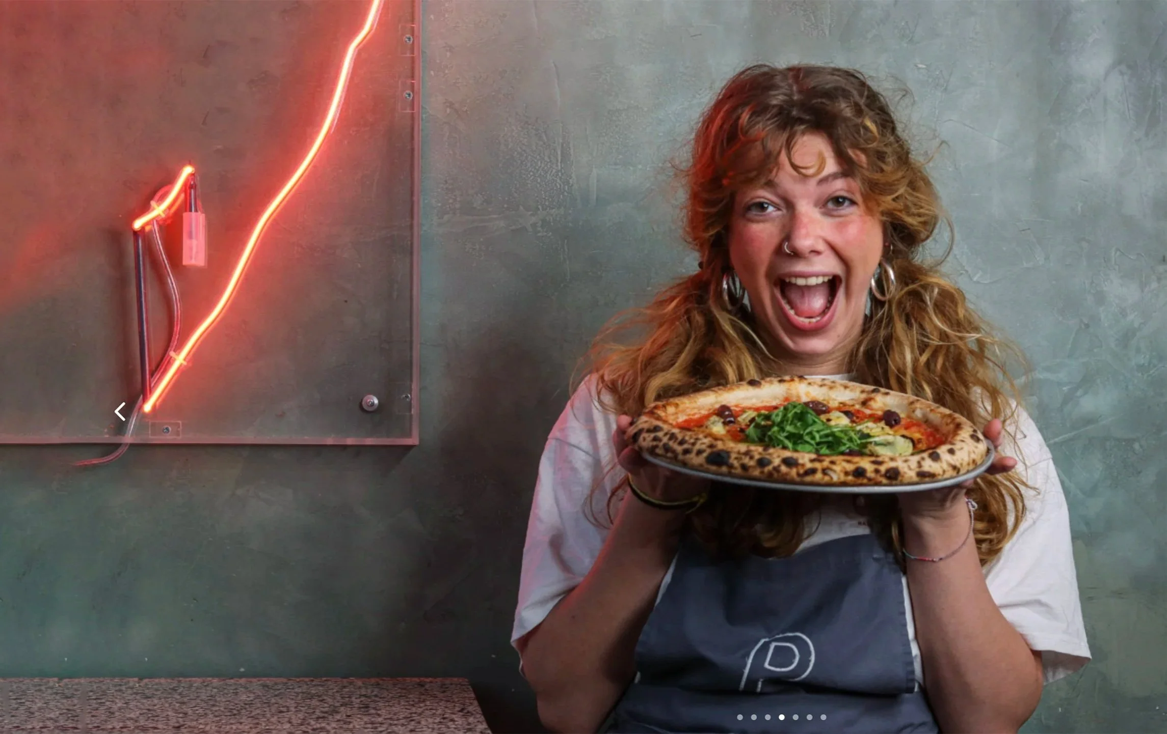 A woman with red hair, wearing a nose ring and earrings, is holding a large pizza with a crispy crust and various toppings, smiling excitedly in front of a gray wall with a neon light.