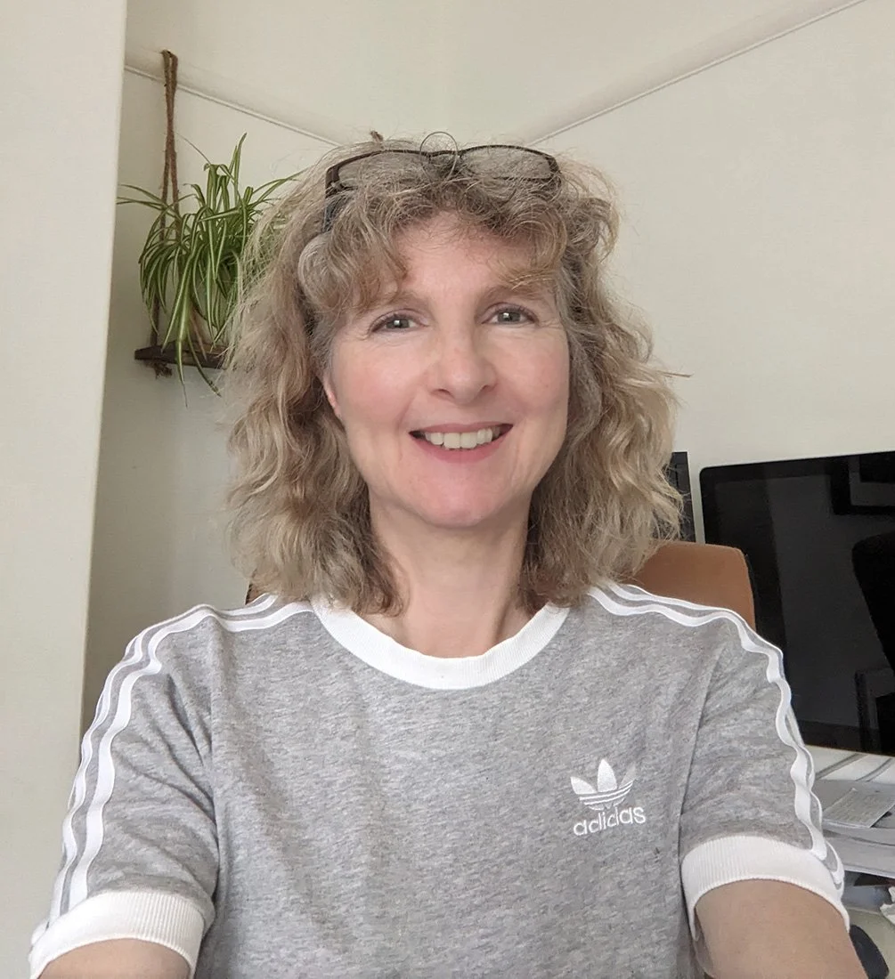 A woman with curly blonde hair, wearing glasses on her head and a grey Adidas t-shirt, smiling at the camera indoors with a computer and a hanging plant in the background.