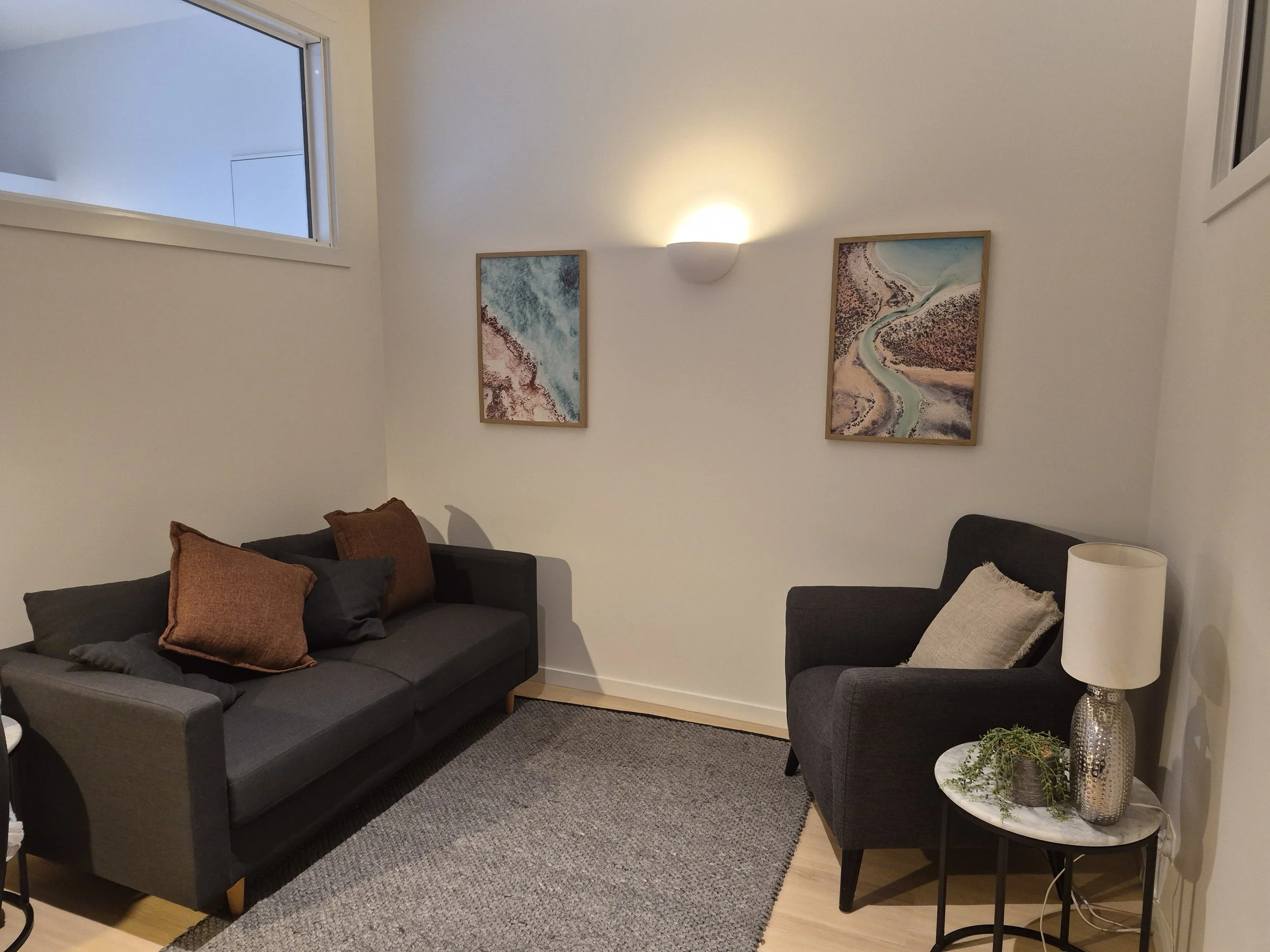 A cozy consulting room with a dark grey sofa, armchair, and landscape prints on the wall.