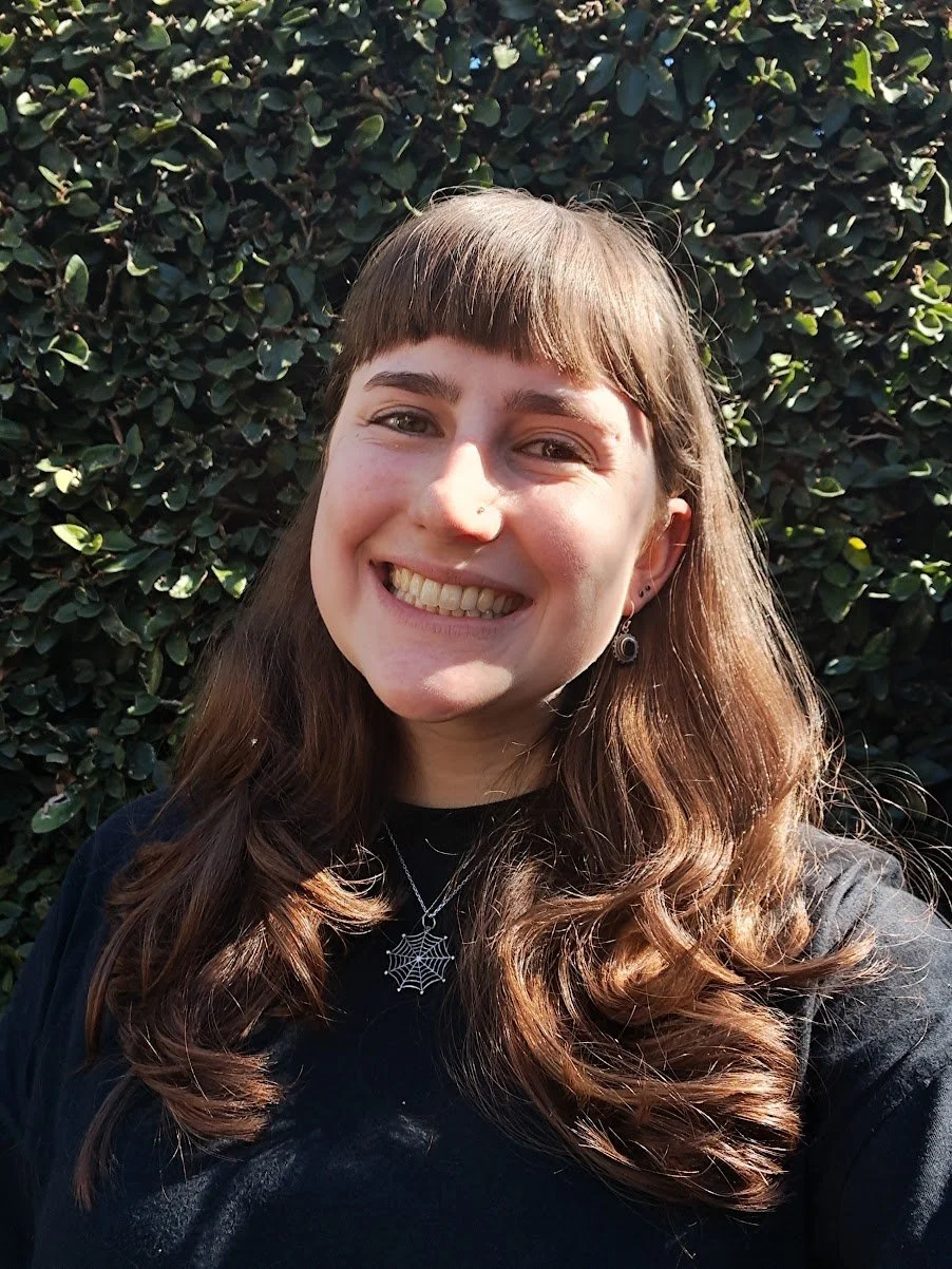 A woman with long brown hair and bangs smiling outdoors in front of a leafy hedge, wearing a black top, a spiderweb necklace, and earrings.
