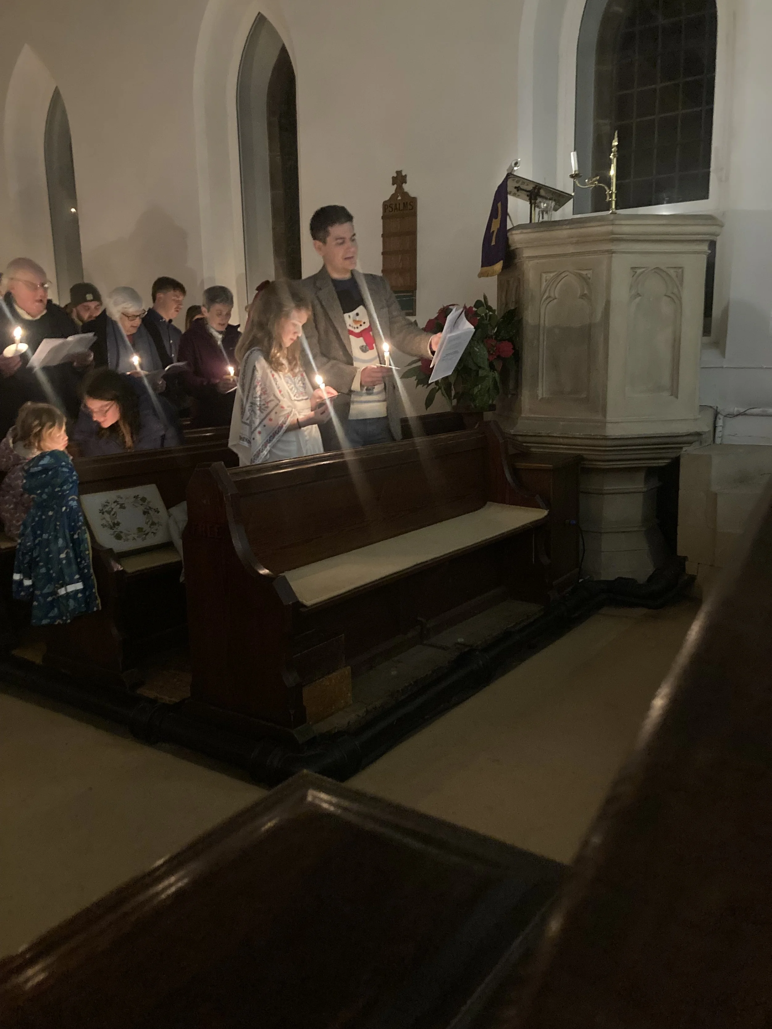A group of people participating in a candlelight service inside Jesus Church Troutbeck, standing in the pews and singing a carol.