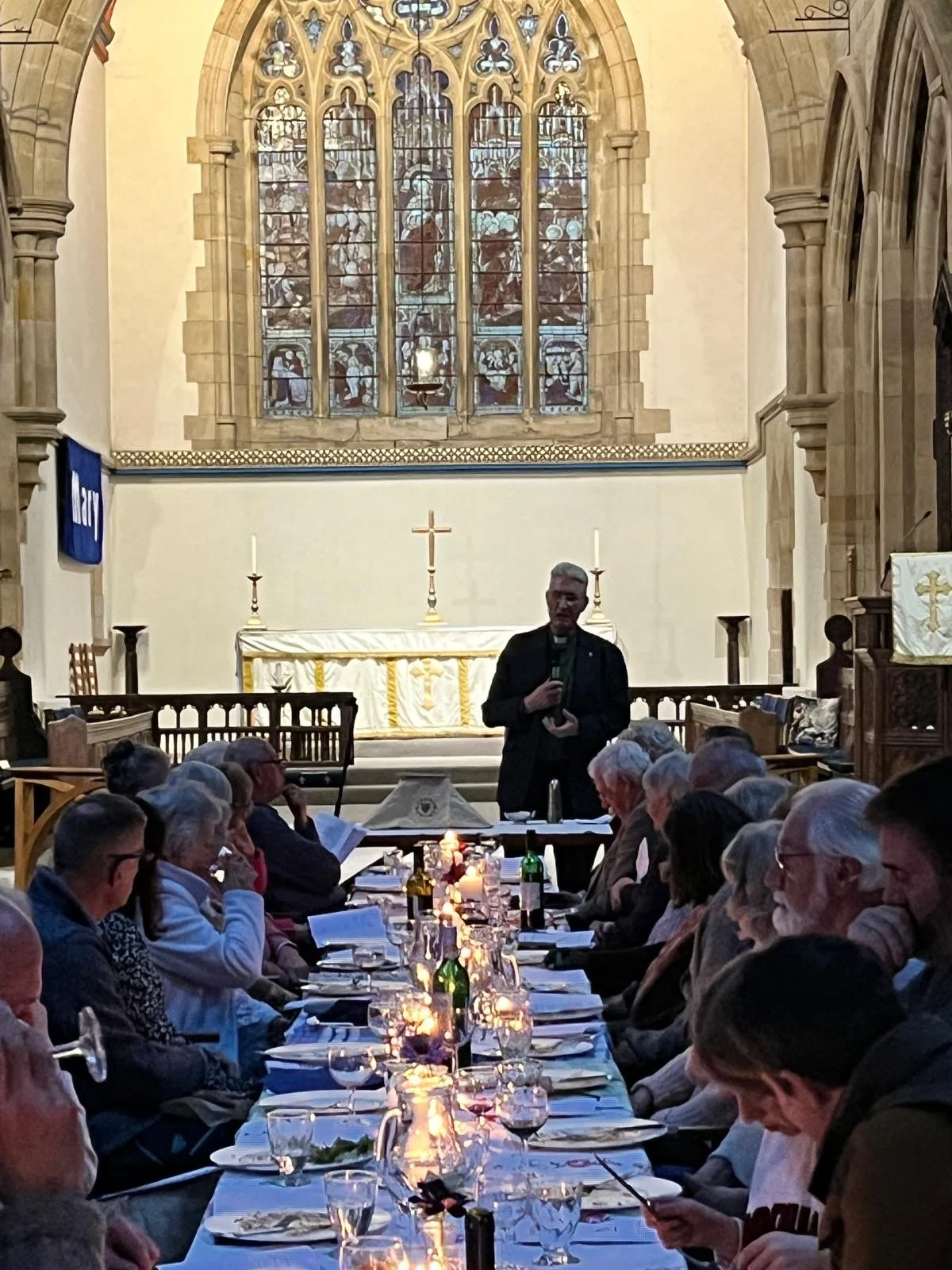 A large group of people seated at a long dining table inside a church, with a priest standing and speaking in front of a congregation, illuminated by candles, with an altar and stained glass window behind.
