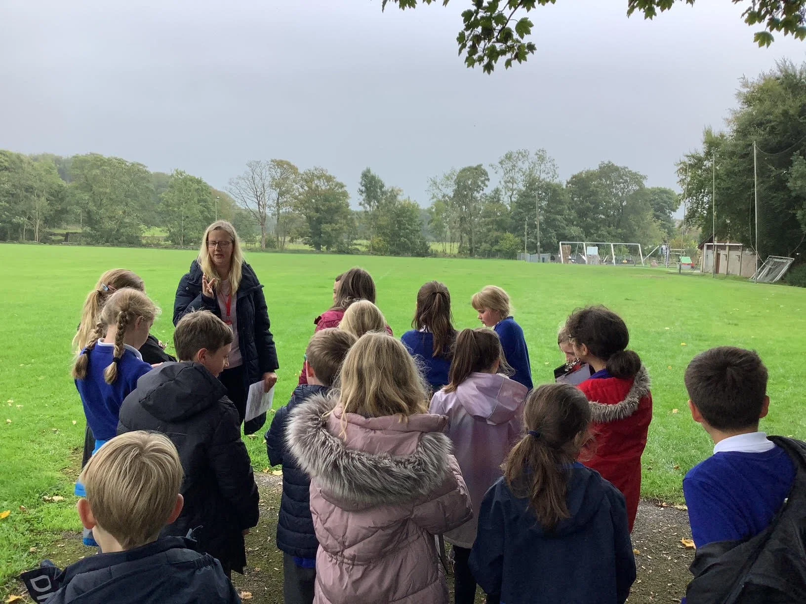 A group of children and a woman standing outdoors on a grassy field, possibly on a school trip or outdoor activity, with trees and sports equipment in the background under an overcast sky.