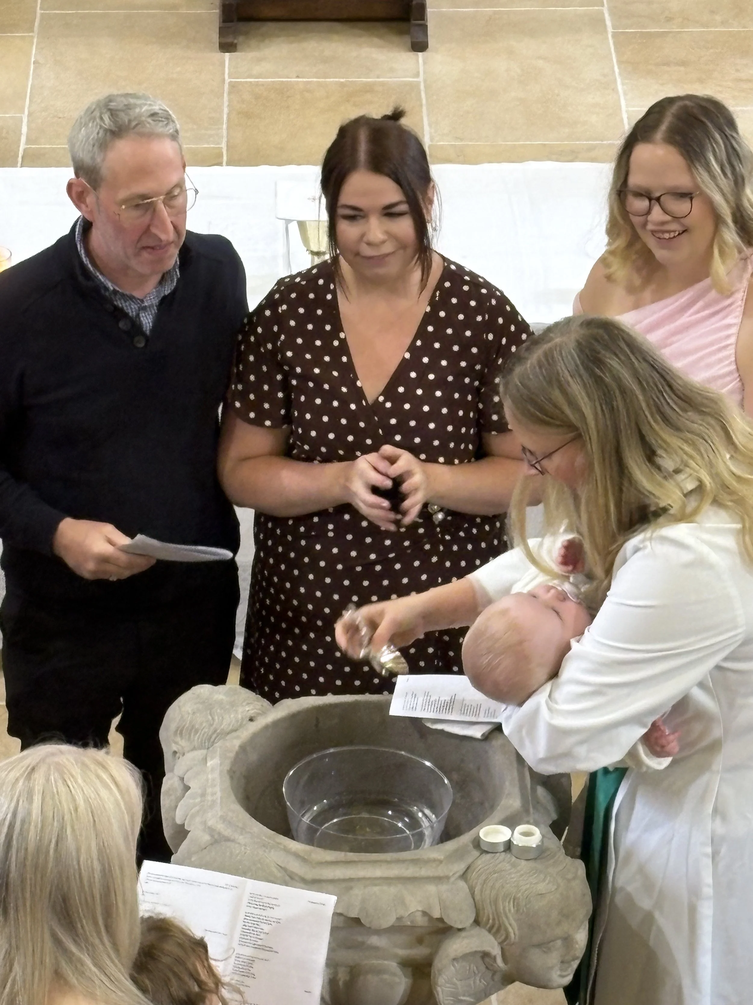 A group of people gathered around a stone baptismal font, with Rev. Olivia Haines holding a baby, preparing for a baptism ceremony in a St Anne's Church, Ings.