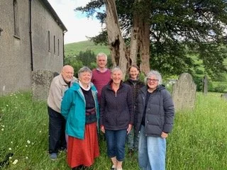Group of seven people standing outside St Cuthbert's Church Kentmere on grass near large trees, smiling at the camera.