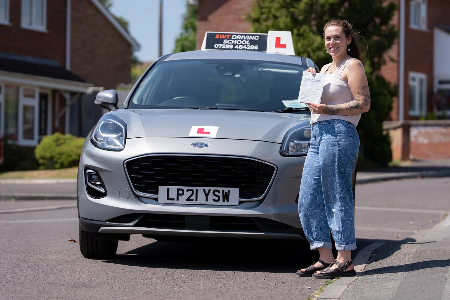 A smiling woman standing on a street in Trowbridge next to a silver Ford Puma holding a driving test pass certificate. The car has a rooftop sign with the text 'SWT DRIVING SCHOOL' and a red 'L' learner driver sign on the bonnet.